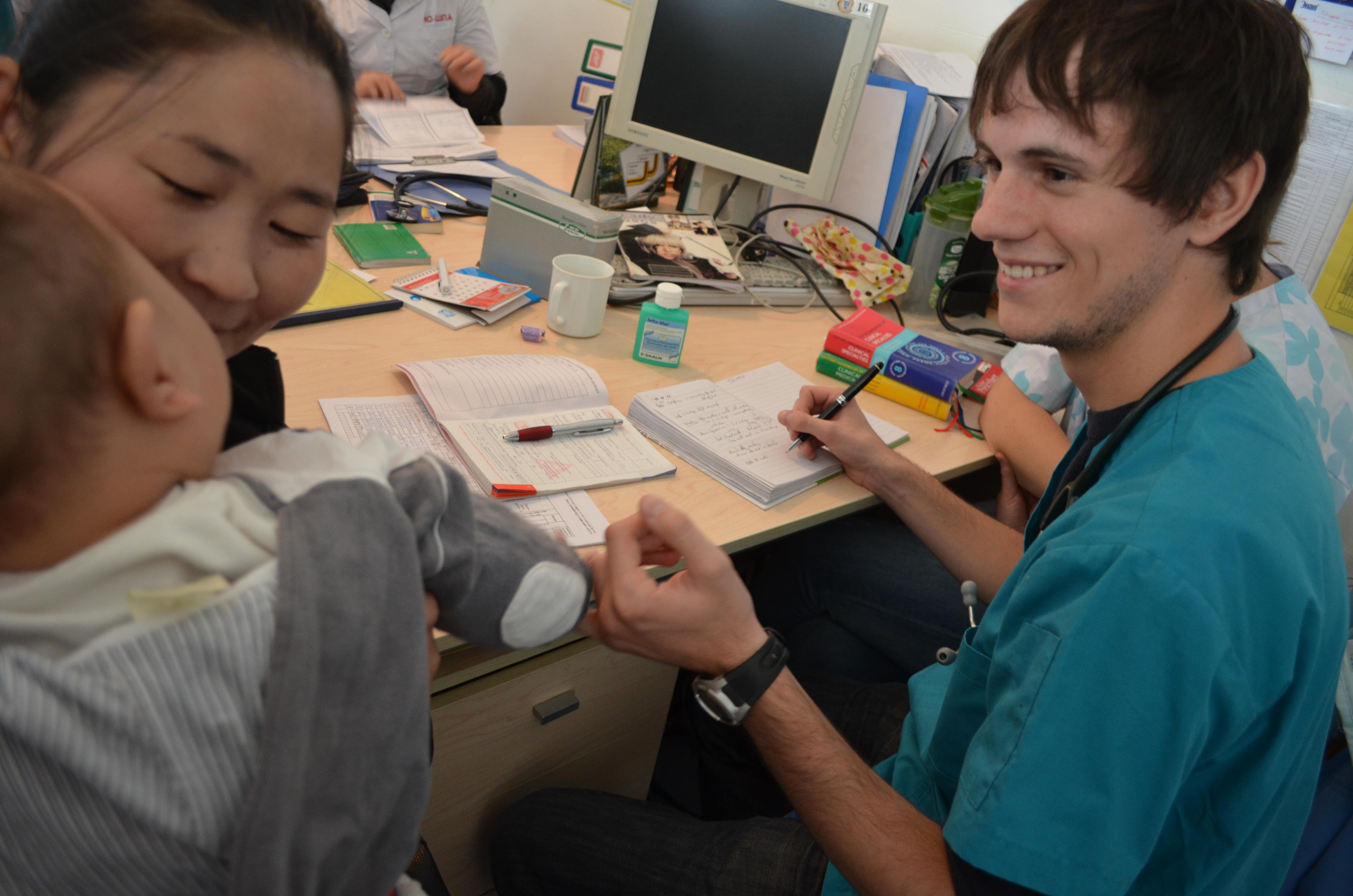Projects Abroad medical intern takes notes on a mother and her child at medical examination as part of a Medicine Internship in Mongolia. Male medical intern sits down with a local woman and her baby during an examination held in Mongolia.